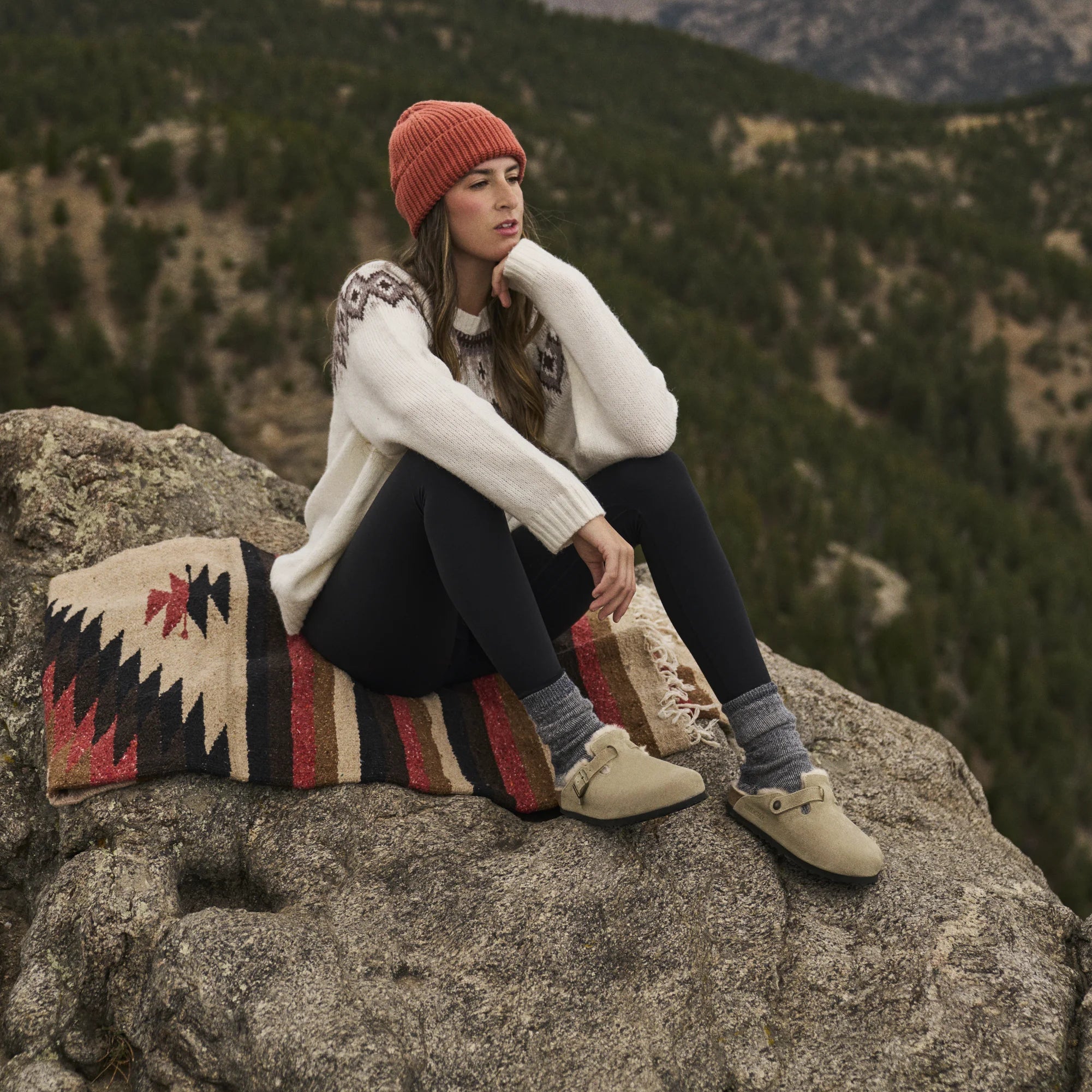 A woman sits on a large rock overlooking a mountain landscape, wearing a white knit sweater, dark leggings, beige boots, and a rust-orange beanie. A patterned blanket rests beside her.