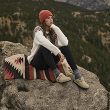 A woman sits on a large rock overlooking a mountain landscape, wearing a white knit sweater, dark leggings, beige boots, and a rust-orange beanie. A patterned blanket rests beside her.