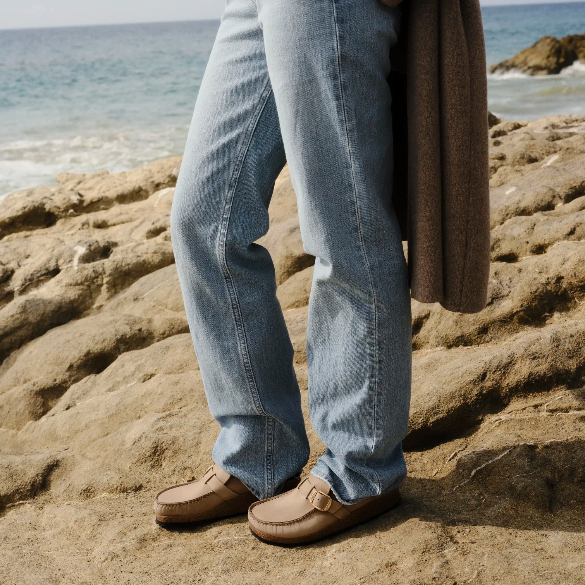 A person standing on rocky beach ground wearing light-wash blue jeans, a blue sweater, a brown cardigan, and brown loafers, with the ocean and sky in the background.