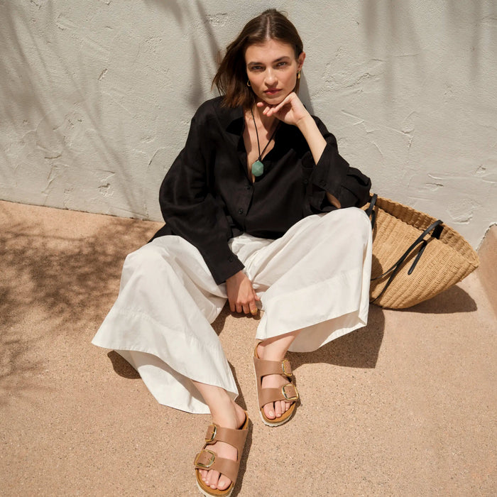 A woman sits on a concrete ledge against a textured white wall, wearing a black button-up shirt, white wide-leg pants, and brown slide sandals. A woven straw tote bag rests beside her.