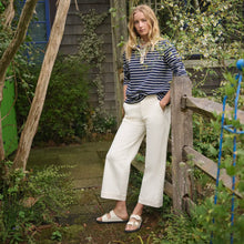 Woman standing in a garden wearing a striped shirt and white pants and ecru sandals
