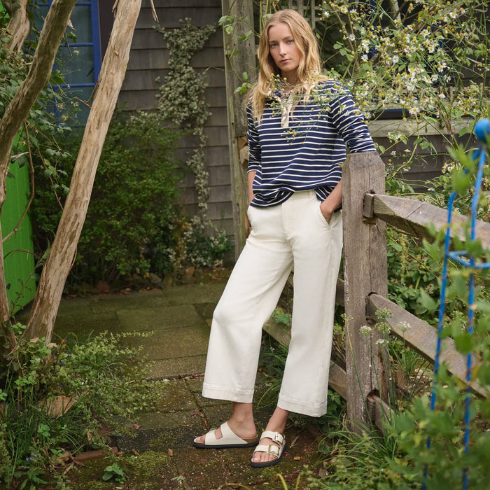 Woman standing in a garden wearing a striped shirt and white pants and ecru sandals