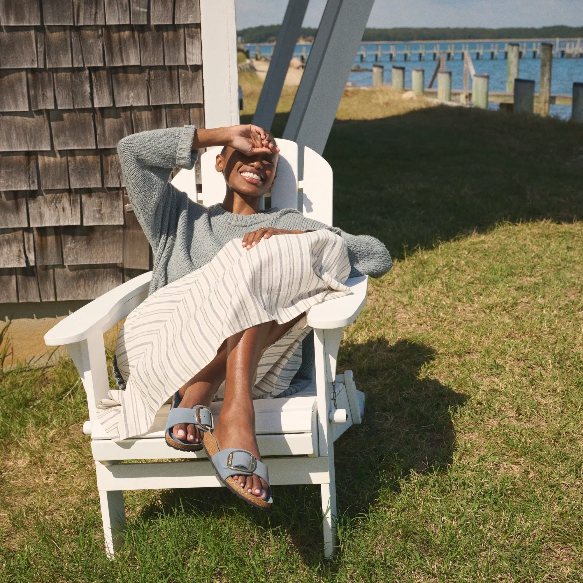 Person lounging on a white Adirondack chair by a wooden cabin with a view of the water.