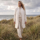 A woman stands in a grassy coastal dune wearing a long cream quilted zip-front coat over a matching cream knit top and trousers, with brown shoes, under a cloudy sky with the sea in the background.