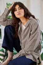 A woman wearing an oversized brown and beige vertically striped button-up shirt, dark blue jeans, and a white undershirt, sitting on a green and white striped couch in a bright room.