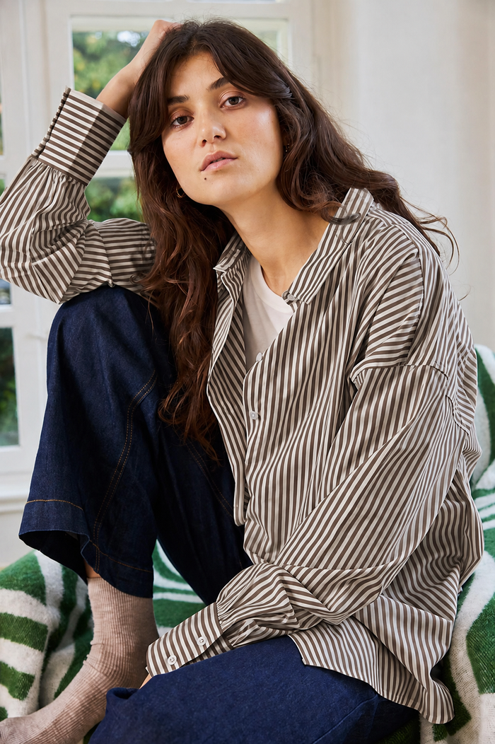 A woman wearing an oversized brown and beige vertically striped button-up shirt, dark blue jeans, and a white undershirt, sitting on a green and white striped couch in a bright room.