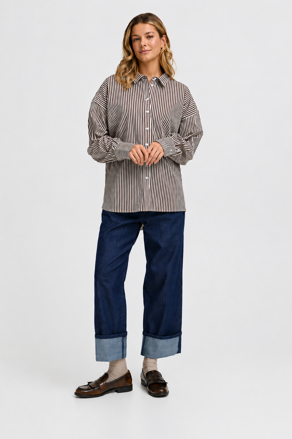 A woman modeling a long-sleeve button-up shirt with thin vertical stripes in brown and beige, paired with dark blue cuffed jeans and brown loafers, against a plain light gray studio background.