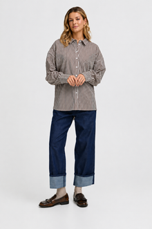 A woman modeling a long-sleeve button-up shirt with thin vertical stripes in brown and beige, paired with dark blue cuffed jeans and brown loafers, against a plain light gray studio background.