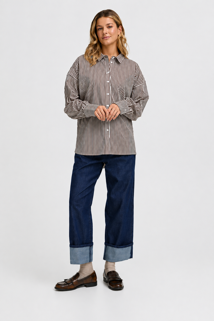 A woman modeling a long-sleeve button-up shirt with thin vertical stripes in brown and beige, paired with dark blue cuffed jeans and brown loafers, against a plain light gray studio background.