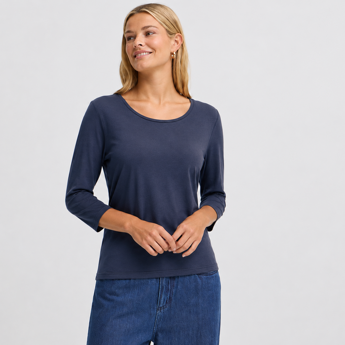 A woman modeling a navy blue three-quarter sleeve top with a wide scoop neckline, paired with blue jeans, against a light gray studio background.