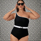 Woman wearing a black one-shoulder swimsuit with a white belt against a geometric patterned wall.