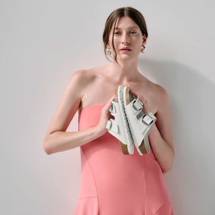 Woman in a pink dress holding white sandals against a plain background