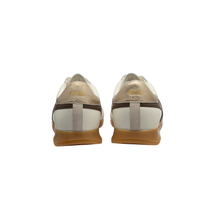 A pair of low-top sneakers shown from the back, featuring metallic gold and off-white leather uppers with dark brown stripes and gum soles.